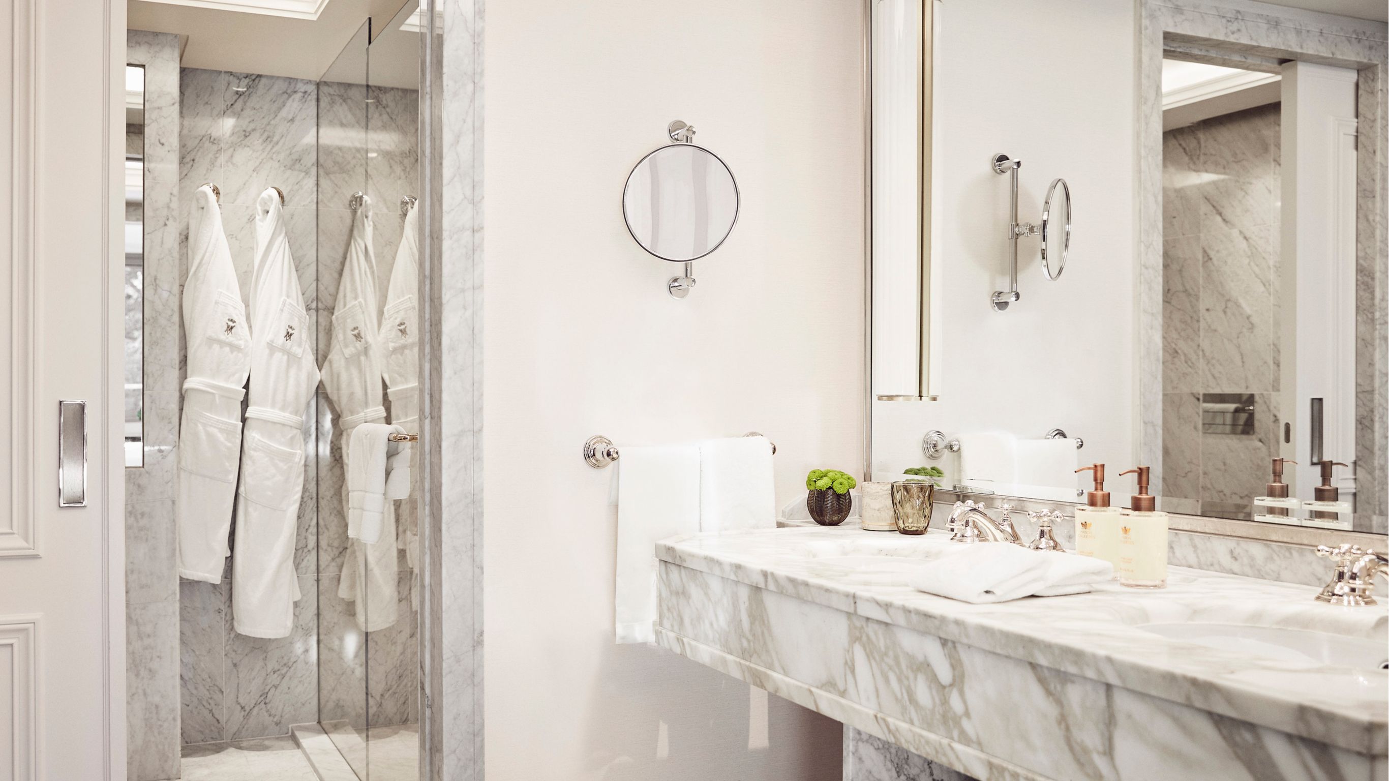A hotel bathroom features white robes hanging on the wall, with a large mirror reflecting the space.